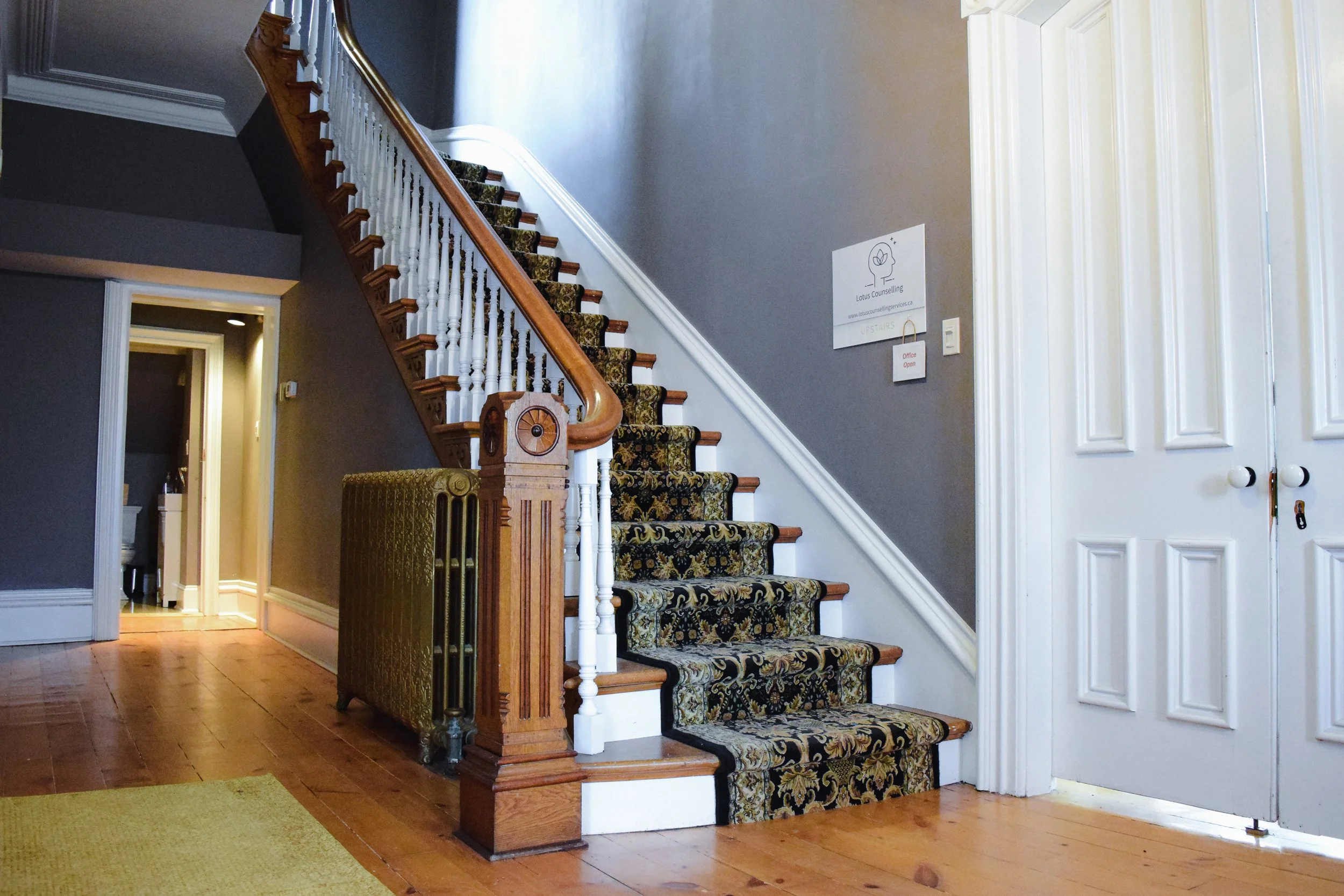 The entryway at Lotus Counselling Services, a bright foyer with a wooden staircase, patterned runner, and an antique cast-iron radiator